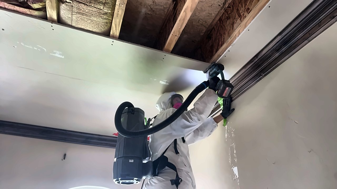 A 4D Restoration tech in a Tyvek suit and respirator works an opened ceiling with a vacuum-captured power tool, joists and insulation exposed above.