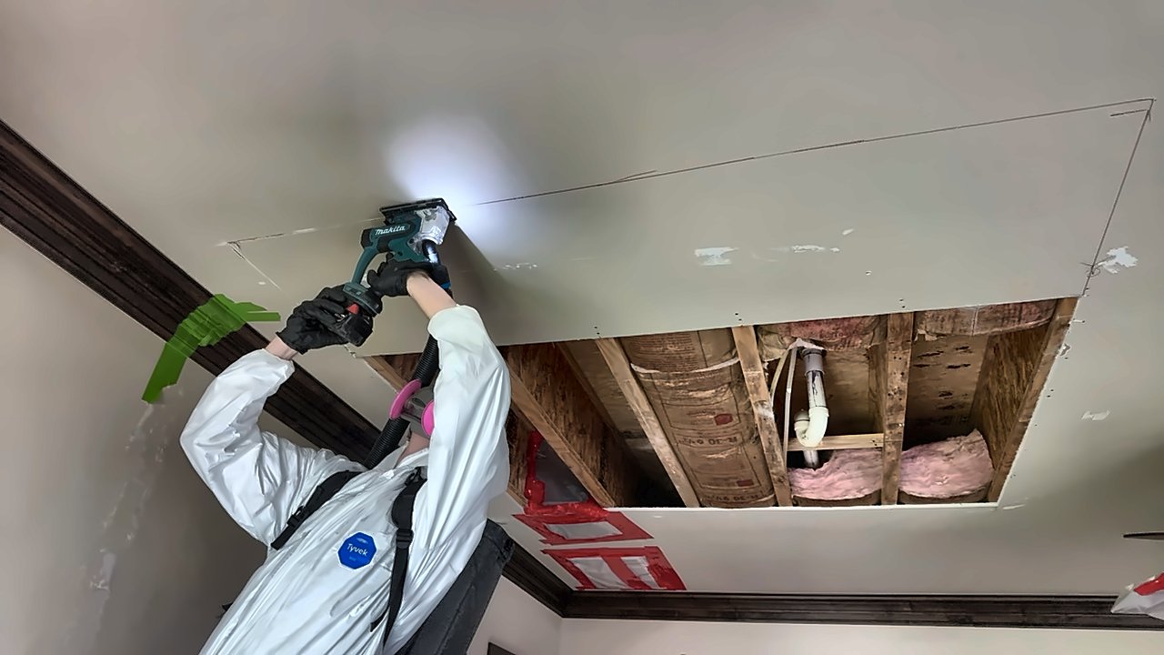 A 4D Restoration tech in a Tyvek suit and respirator cuts into an opened ceiling with a vacuum-captured Makita tool, exposing joists and insulation overhead.