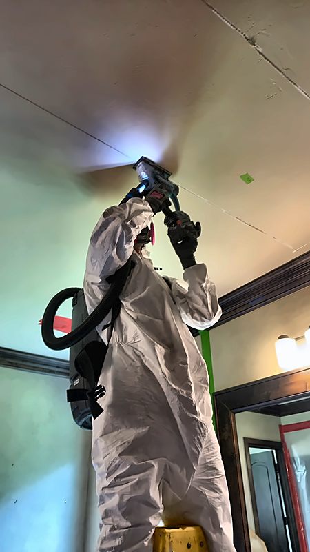 A suited 4D Restoration tech on a ladder works a ceiling with a handheld power tool.