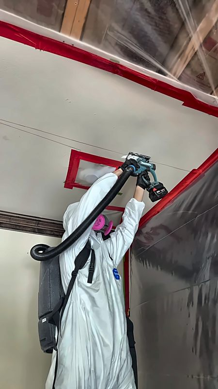 A 4D Restoration tech in a full protective suit and respirator cuts into a ceiling with a vacuum-captured power tool.